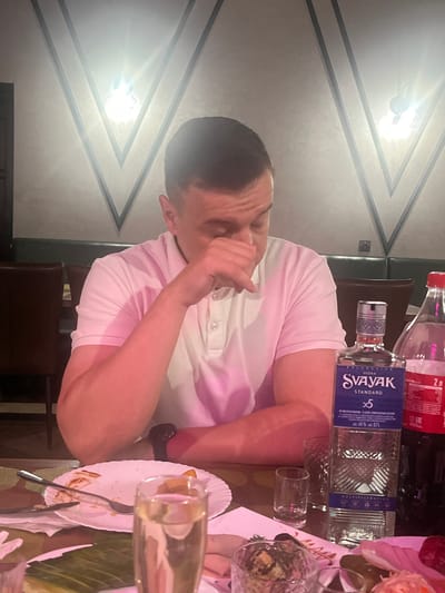 In a dimly lit restaurant with a textured ceiling featuring V-shaped light fixtures, a man in a pink polo shirt sits at a table, appearing distressed as he covers his face with his hand. On the table, a partially eaten meal on a white plate, a fork, a glass of champagne, a bottle of "SVAYAK STANDARD" vodka, and a two-liter bottle of soda are visible. The overall ambiance suggests a somber or unpleasant dining experience.