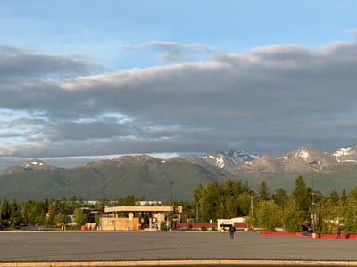 This image depicts an expansive asphalt parking lot in Anchorage, Alaska, with a mountain range serving as a dramatic backdrop. The parking lot appears largely empty, with faint tire marks suggesting recent use. In the mid-ground, a small gas station with pumps and a convenience store is visible, accompanied by a Burger King restaurant. Scattered trees and vegetation dot the landscape between the parking lot and the mountains. The sky is partly cloudy, with a mix of blue sky and grey clouds illuminated by the soft light of either dawn or dusk. Two individuals are present in the parking lot; one walks towards the left side of the frame, and another walks towards the right. The overall scene suggests a commercial area on the outskirts of the city, with natural scenery prominent in the background.