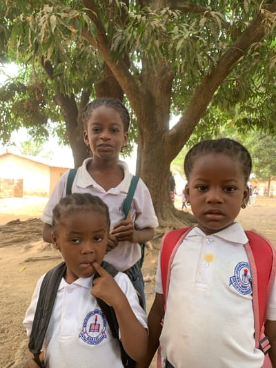 Three Black children, appearing to be girls, are photographed outdoors in what is identified as Garaku, Nigeria. The children are positioned in the foreground against a backdrop featuring a large tree and sparse buildings. All three wear white short-sleeved polo shirts, indicative of school uniforms, each bearing a circular blue and white emblem on the left chest with text, partially visible, identifying it as "ADULT LITERACY SCHOOL GARAKU" and "The Fear of God Is the Beginning of Wisdom."
The central child, positioned slightly behind the other two, has braided hair and a faint smile, looking directly at the camera. She wears dark bottoms and green backpack straps are visible over her shoulders. To her lower left, another child, also with braided hair, has her right index finger resting on her lower lip and her gaze directed slightly downward and to the left. She wears dark bottoms, and black backpack straps are visible. To the right, the third child, with braided hair, maintains a serious expression and looks directly forward. She wears a red backpack and her white polo shirt displays a distinct yellow stain on the upper right chest and additional dirt marks on the lower front.
The immediate ground beneath the children is dry, light-brown earth with exposed tree roots visible in the middle ground. The background is dominated by a large, leafy tree with a thick trunk and numerous branches. To the far left, a light-colored building with a dark roof and exposed brickwork is visible. Further in the background on the right, blurry figures of other individuals are discernible, suggesting a communal or schoolyard setting. The scene is illuminated by natural daylight.