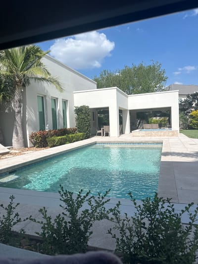 This image depicts a modern residential backyard in McAllen, United States of America, under a bright, sunny sky with scattered white cumulus clouds. A rectangular swimming pool with clear, turquoise-blue water occupies the mid-ground, featuring light blue and dark blue mosaic tiling around its perimeter and light-colored stone coping. Steps are visible on the pool's left side.
To the left, a contemporary white stucco building, likely a house, exhibits a flat roofline and several tall, vertical, dark-framed windows. A large palm tree stands prominently beside this structure, with a landscaped bed containing red and green foliage and neatly trimmed green hedges along its base.
In the center-right, a series of connected, open-air white stucco structures with flat roofs form covered patio areas. One section houses a light-colored outdoor dining table with four chairs. A further section contains lounge furniture and an elevated, tiled spa or small pool. Light-colored paving stones form the deck around the pool and extend to these structures.
Green grass lawns and various trees are visible in the background. The immediate foreground is partially obscured by out-of-focus green leafy bushes and an indistinct dark object at the very bottom of the frame. The scene is devoid of any individuals, actions, or interactions.