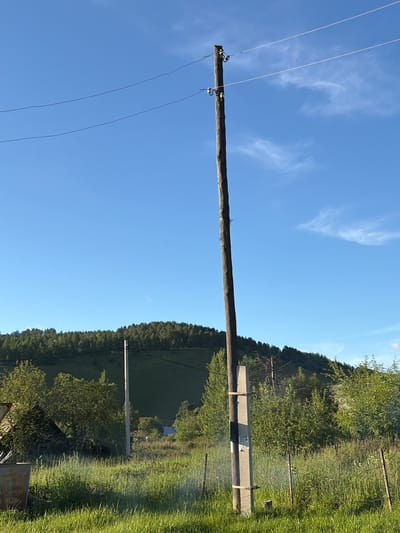 A low-angle shot captures a wooden utility pole in the foreground, supporting overhead wires against a clear blue sky with wispy clouds. The pole has insulators and wires attached near its top. To the right of the pole is a concrete base. In the midground, a dense, green forest blankets a hill under the sky. A rural landscape unfolds with tall, green grass in the foreground, interspersed with smaller trees and bushes. A rustic building, possibly a shed or old house, is visible to the left, partially obscured by vegetation. In the distance, a single, lighter colored utility pole stands among the trees. The scene depicts a tranquil, outdoor setting, likely in a rural or semi-rural area. The geographical location is identified as Skovorodino, Russia.