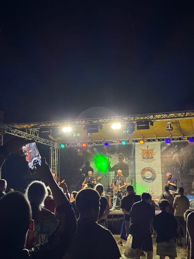 This image depicts an outdoor music concert at night, likely part of the "July Morning Tutrakan" festival, as indicated by the banner visible on the stage. The stage is equipped with lighting and sound systems, and a band is performing. Several people are visible in the foreground, some holding up phones to record or photograph the performance. The background is dark, suggesting it is nighttime, and there are subtle indications of falling rain or debris in the air. The overall atmosphere appears to be that of a lively event. The location is identified as Tutrakan, Bulgaria.