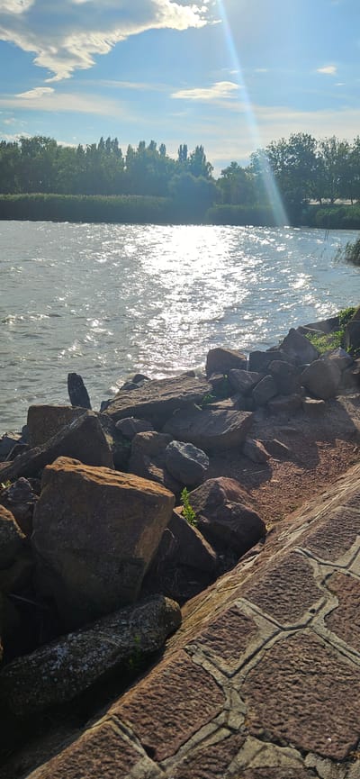The image depicts a sun-drenched waterfront scene with a rocky embankment and a calm body of water. The sky is a vibrant blue with scattered white clouds, and the sun's rays create a sparkling effect on the water's surface. The embankment is covered with large, rough stones, some of which are reddish-brown and others are darker gray. Green vegetation, including small plants and grasses, grows between the rocks. In the foreground, a paved walkway with patterned stones is visible on the right side. The opposite shore features a dense line of trees and vegetation. The image is taken from a low angle, emphasizing the texture of the rocks and the expanse of the water. There are no people or animals visible in the frame. The setting suggests a natural or semi-natural waterfront environment, possibly a park or a recreational area. Balatongyörök is a village located on the northern shore of Lake Balaton in Hungary, known for its scenic beauty and waterfront access, which aligns with the visual elements present in the photograph.