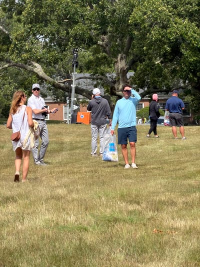 In the Town of Southampton, United States of America, a group of people are gathered on a grassy field. The image captures a sunny day with a large tree providing shade.
In the foreground, a woman in a white dress with a brown purse and a light-colored scarf is walking away from the camera. Beside her, a man in a white polo shirt and gray pants is holding a phone in his left hand and a dark object in his right. He is wearing sunglasses and a white cap.
Further back, another man in a gray hoodie and light gray pants is holding a clear plastic bag filled with items. To his right, a man in a light blue long-sleeved shirt and dark blue shorts is looking up and shielding his eyes from the sun. He is also holding a clear plastic bag.
In the background, a woman in a black jacket and pink head covering is walking, and a man in a navy blue shirt and shorts is standing. An orange structure with a "W" logo is visible behind the man in the white polo shirt. The environment is a grassy field with dry patches, and a large tree with lush green leaves dominates the upper portion of the image. The sky is a clear blue.