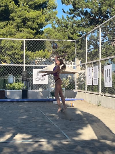 The image depicts a beach volleyball match in progress on a sand court. A female athlete, in mid-air, is in the process of serving the ball. She is wearing a purple bikini. The court is enclosed by a fence with trees and buildings visible in the background. The location is specified as Gošići, Montenegro. The overall scene is one of outdoor athletic activity on a sunny day.