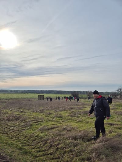An outdoor scene in Stara Reka, Bulgaria, depicts a group of individuals traversing an open field under a partly cloudy sky. The bright, diffused sun is visible in the upper left quadrant. The ground consists of uneven terrain covered with patches of green grass interspersed with significant areas of dry, light brown vegetation. In the foreground, positioned to the right, a man walks from left to right, his body angled slightly towards the camera. He wears a black beanie, a black padded jacket with a red inner lining, black pants, and dark footwear. He carries a dark backpack and holds a white plastic bottle in his right hand, looking forward with a slight smile. In the midground, a group of approximately 20-30 people, many dressed in colorful jackets (red, blue, green), walk across the field from left to right. A dark, rectangular, box-like structure stands isolated in the field to the left of the main group. In the distant midground, a small flag, possibly Bulgarian, is visible held by one of the individuals. A dense treeline or forest forms the horizon line, separating the field from a greener expanse of land beyond.