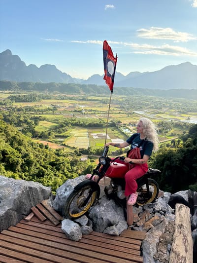 In Hong Ngern Village, Laos, a blonde woman with curly hair is seated on a red motorcycle. She is wearing a dark t-shirt and bright red pants, with pink Crocs on her feet. A red bandana is tied around her neck, and she has tattoos on her arms. The motorcycle is positioned on a rocky outcrop overlooking a valley filled with lush green vegetation and rice paddies. A flagpole with the flag of Laos stands behind the motorcycle, waving in the breeze. The landscape is characterized by rolling hills and majestic, mist-shrouded mountains in the distance, bathed in the soft light of a late afternoon sun. Wooden planks are laid out on the rocks, forming a makeshift platform.