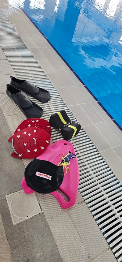 The scene depicts a collection of swimming equipment neatly arranged on the deck of an indoor swimming pool in Umm Salal Ali, Qatar. The pool features calm, blue water with reflections of an overhead structure, bordered by light-colored, textured tiles and a dark blue dividing line. White, slatted overflow grates run parallel to the pool edge, separating the water from the main deck area.

The swimming equipment, from top to bottom and left to right in the image, includes:
*   Two black swimming fins (flippers) placed side-by-side on the grates.
*   Two red hand paddles, adorned with multiple white stars and small perforations, are stacked next to the fins.
*   A black pull buoy with a yellow strap bearing the brand name "NABAIJI" is positioned near the hand paddles.
*   A pink kickboard is situated below the hand paddles and pull buoy.
*   A black swim cap, featuring a red and white "Yakult" logo, rests on the pink kickboard.
*   A pair of blue and yellow swimming goggles with clear lenses is draped over the kickboard.

The deck consists of light grey/beige square tiles with a subtle textured pattern. Further away from the pool, darker grey, rough-textured tiles are visible, one of which contains a circular white grate. No individuals are present in the image.