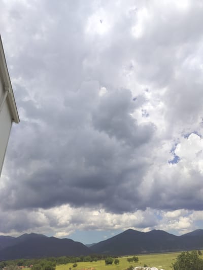 A wide shot captures a stormy sky over a rural landscape in Gorno Sahrane, Bulgaria. Dark, heavy clouds dominate the upper portion of the frame, with lighter, scattered clouds and patches of blue sky visible higher up. In the foreground, the white eaves and downspout of a building are visible on the left edge. Below the sky, a range of dark, silhouetted mountains forms the horizon. In front of the mountains, a green, open field stretches across the midground, dotted with scattered trees. Towards the left of the field, several houses with red roofs are visible among the trees. The overall scene depicts an overcast, possibly pre-storm, atmospheric condition over a pastoral setting.