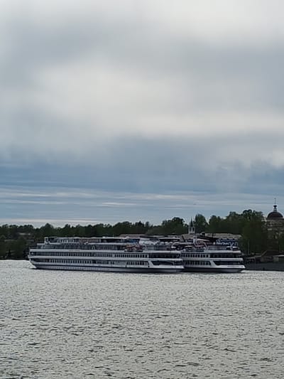 The image depicts a long, white passenger river cruise ship with multiple decks, docked near the shoreline of the Volga River in Myshkin, Russia. The ship is the primary subject and appears to be several stories high, with numerous windows. A line of trees and building domes are visible on the shore in the background, and the sky is overcast with a mix of gray and white clouds. The water of the Volga River occupies the lower portion of the image, showing a slight ripple and texture.