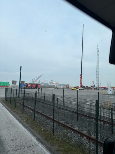 The scene depicts an industrial port area at Maasvlakte, Rotterdam, Netherlands, under an overcast sky. The image is captured from within a vehicle, with the top edge of a windshield visible. In the immediate foreground, a grey asphalt surface with a white line runs along the bottom left. A dark green metal mesh fence extends horizontally across the frame, behind which two sets of railway tracks are laid on gravel ballast.

Beyond the tracks and fence, a large paved industrial yard unfolds. Several vehicles are parked or positioned within this area. From left to right, a green tarped semi-trailer truck is visible, followed by multiple semi-trailer trucks, some with red containers marked "ONE". A yellow utility vehicle, possibly an ambulance or service van, is parked next to a white sedan. Near these vehicles, at least two individuals are present, one appearing to wear an orange high-visibility vest.

A prominent, tall red mobile crane with its boom fully extended vertically dominates the central midground. To its right, a tall metal lattice tower stands. Various shipping containers and other industrial equipment are scattered across the paved yard. In the distant background, a large cargo ship or tanker is docked, surrounded by several red gantry cranes, indicative of active port operations. The sky is uniformly light grey and overcast.