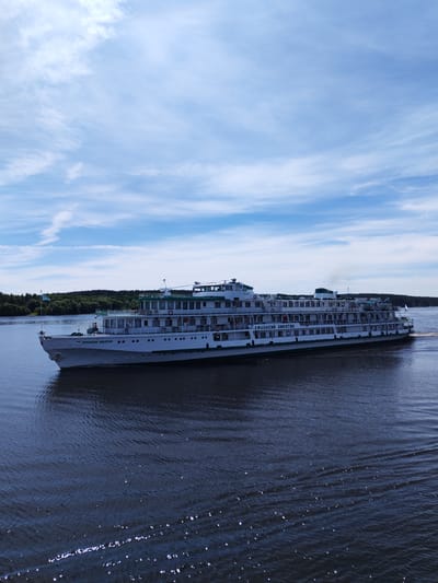 The image displays a large, multi-deck passenger ship, identified by the Cyrillic lettering "АФАНАСИЙ НИКИТИН" (Afanasiy Nikitin) on its bow and port side. The vessel is predominantly white with green trim along its decks and railings, and features numerous windows for cabins, open deck spaces, and visible lifeboats. A light blue flag with an emblem flies from the bow, and a small white flag is positioned at the stern. The ship is in motion on a dark blue body of water, creating a discernible wake behind it. Subtle exhaust smoke is visible from a funnel located on the upper deck towards the stern. Reflections of sunlight are present on the water's surface, particularly in the foreground. In the background, a dense, green forested shoreline extends along the distant bank of the waterway. The sky above is light blue with scattered white cirrus and cumulus clouds. Several individuals are faintly visible on the ship's decks. This scene is located in the vicinity of Nosovo, Russia.