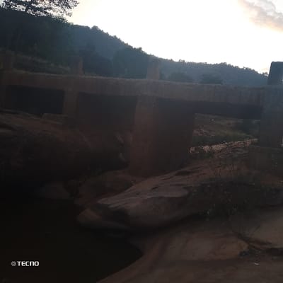 A rugged landscape features a weathered, concrete bridge spanning a rocky, shallow stream. The bridge has multiple openings, supported by sturdy pillars. The setting sun casts warm tones across the scene, illuminating the uneven rock formations and sparse vegetation. A distant, tree-covered hill rises in the background beneath a cloudy sky. The overall impression is one of a natural, slightly desolate, but sturdy environment.