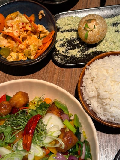 A top-down shot shows a table with several dishes. At the top is a brown bowl filled with white rice. To the left is a small, black rectangular plate with a round, golden-brown puff pastry topped with green sprouts, sitting on a bed of green powder. Below this is a small, blue bowl containing a salad of shredded cabbage, carrots, and green peppers. To the right is a beige oval plate filled with stir-fried vegetables, including broccoli, red and yellow peppers, red onions, and pieces of glazed chicken. A whole red chili pepper and fresh green herbs garnish the dish. The table is made of light-brown wood. The location is Kiryat Ono, Israel.