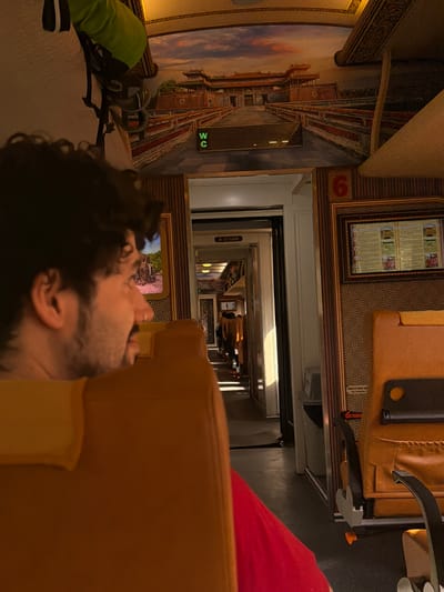 The image shows the interior of a train traveling in Huế, Vietnam. The photo captures a man reclining in a seat, with the train's corridor visible in the background. The train car walls are decorated with images, and a sign indicates the restroom is nearby.