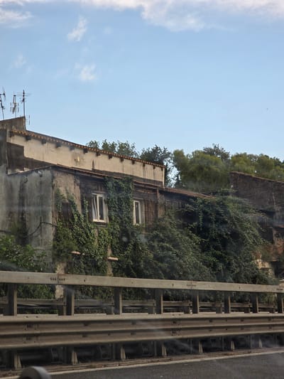 The image shows an old, weathered building partially covered in ivy, viewed from behind a metal guardrail on what appears to be a road or highway. The building has at least two visible windows with white frames. The sky above is light blue with scattered white clouds. In the foreground, the metallic guardrail dominates the lower portion of the image. The overall impression is one of an urban or semi-urban setting with signs of age and neglect, typical of older structures in a city like Salerno, Italy.