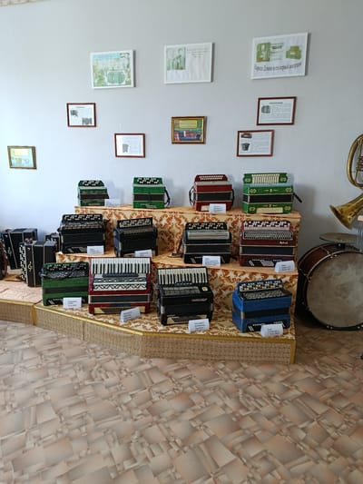 The image shows a museum display of accordions. The accordions are arranged on tiered platforms covered with patterned fabric. There are framed pictures and documents displayed on the wall behind the accordions. To the right of the accordions, part of a large drum and a brass instrument are visible. The floor is covered with a patterned linoleum. The context provided indicates the location is likely the Смілавіцкі сельскі Савет in Belarus. There are no people visible in the image.