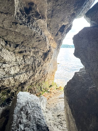 The image shows a view from inside a cave or rock overhang, looking out towards the sea. The foreground is dominated by large, rough-textured rocks, which create a natural frame for the scene. Some patches of green moss or algae are visible on the rock surfaces in the shaded areas. Beyond the rocks, a body of water, likely the sea, reflects the light, with small waves rippling its surface. In the distance, a hazy shoreline with rolling hills and trees can be seen under a bright sky. The location is identified as Dom Otdykha Zolotoy Plyazh, Russia.