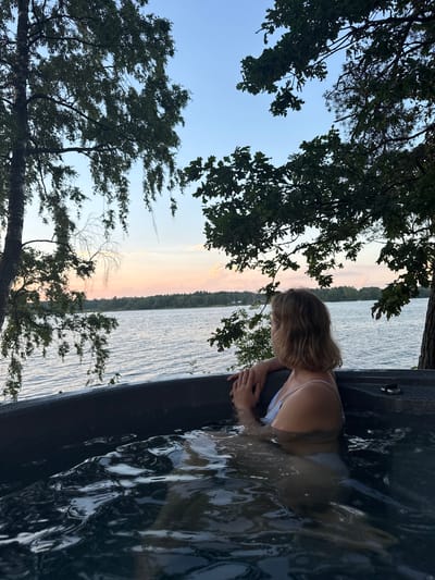 A woman relaxes in a hot tub overlooking a calm lake during twilight. She is partially submerged in the dark water, wearing a light-colored bikini. Her blonde hair is short, and she is looking out towards the water. The hot tub is dark-colored and positioned on a wooden deck or platform near the water's edge. Trees with green leaves frame the scene, casting shadows and partially obscuring the view of the sky. The sky exhibits a soft gradient of blue and pink hues, characteristic of sunset or sunrise. The location is identified as Priedesmuiža, Latvia.