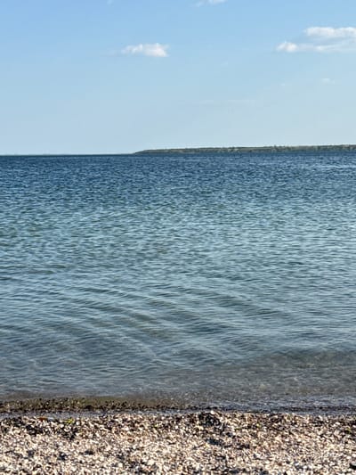 The image depicts a tranquil, natural scene. The foreground is dominated by the edge of a body of water, likely a lake or a wide river, with gentle ripples disturbing its surface. The water transitions from a clearer, lighter shade near the shore to a deeper blue further out. The shoreline is composed of small, light-colored pebbles, creating a textured contrast with the water. In the background, a dense, dark green line of trees or vegetation marks the far bank of the water. The sky above is a pale, clear blue with a few wispy white clouds scattered across it. The lighting suggests it is daytime, likely late morning or early afternoon, given the bright illumination. There is no discernible activity taking place; the scene appears still and serene. The location is identified as Ukrainka, Ukraine. No text is visible in the image.