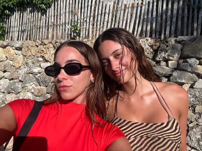The image displays two young adult females photographed outdoors in Cap-d'Ail, France. The female on the left wears black oval sunglasses and a red short-sleeved top, with a black strap visible over her left shoulder. Her dark hair is wet. The female on the right wears a halter-neck top with a light and dark brown horizontal striped pattern; her dark hair also appears wet, and she exhibits a faint smile. Both subjects are positioned closely, facing the camera. The background features a textured stone wall, topped by a weathered wooden fence with vertical slats, partially covered by green foliage. The scene is illuminated by direct sunlight, casting distinct shadows.