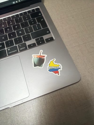 The image shows a close-up, overhead view of a silver laptop with black keys. Two stickers are present on the laptop's surface. One sticker is of a green gourd with a straw, commonly used for drinking mate. The other sticker is a stylized map of Colombia with its flag colors and the word "COLOMBIA" printed on a banner. The laptop is placed on a textured, beige surface, likely a desk or table. The lighting suggests an indoor environment. There is no indication of the city of Cancun, Mexico in the image.