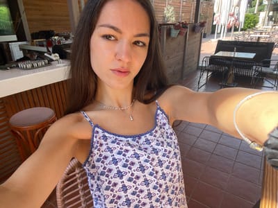 The image appears to be a selfie taken by a woman with long, dark brown hair, brown eyes, and light skin. She is wearing a sleeveless sundress with a white background and a pattern of small purple and blue flowers. The dress has a blue pom-pom trim around the neckline. She is also wearing a delicate silver necklace and a bracelet.
The background shows an outdoor setting. There are dark brown tiled floors, wooden walls, and outdoor furniture including black metal chairs and tables. Umbrellas are visible in the background, suggesting an outdoor cafe or restaurant. There are also plants in pots on a shelf, adding to the ambiance of a Mediterranean outdoor space.
Based on the location (Nea Makri Municipal Unit, Greece), the scene likely depicts a tourist enjoying a day at an outdoor cafe or restaurant in this coastal town. The woman appears relaxed and may be taking a moment to capture a memory of her visit.