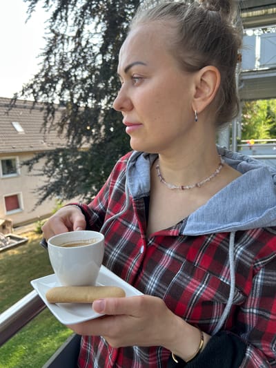 A medium shot captures a woman standing on a balcony, holding a white cup of coffee and a biscuit on a saucer. She is looking to her left, with her hair tied up in a bun. She is wearing a red and black plaid shirt with a grey hood. The background shows a residential building and trees, suggesting an urban environment. The scene is likely set in Velbert, Germany, given the architecture and greenery. The lighting indicates it is daytime, with sunlight filtering through the trees. The woman's posture and the presence of coffee and a biscuit suggest a moment of relaxation or contemplation.