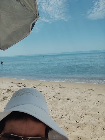 The image shows a person's perspective from under a beach umbrella, looking out at the sea and the sandy shore. The person is wearing a light-colored hat and sunglasses, obscuring most of their face. The umbrella provides shade, indicated by the large fabric structure in the foreground. The beach is sandy with footprints visible, and the sea is calm and blue, with gentle waves lapping at the shore. In the distance, a few people are in the water, and some buoys are scattered across the sea. The sky is clear and blue with scattered white clouds, suggesting a sunny day. The scene evokes a sense of relaxation and enjoyment of a summer day at the beach. The time of day appears to be midday, given the bright sunlight. The location is a beach in Emona, Bulgaria. No text is visible in the image.