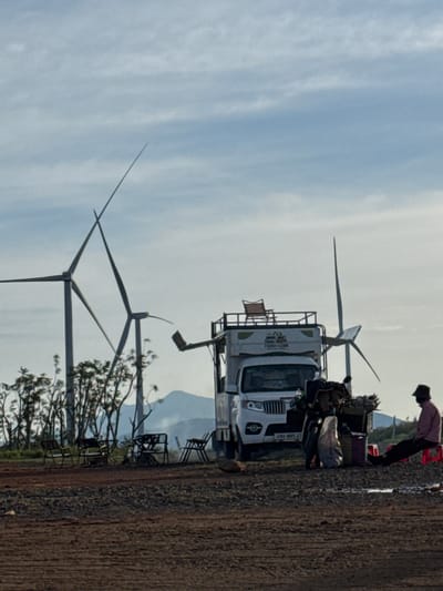 The image depicts an outdoor scene with a cloudy sky and a landscape featuring wind turbines. In the foreground, there is a gravelly and dirt ground. Several metal chairs and tables are set up in the middle ground, arranged in a casual outdoor seating area. Behind the chairs and tables, a white camper van is parked. The van has a rack on its roof with a chair placed on it. A motorcycle is parked next to the van, loaded with various items. To the right of the van, a person wearing a hat and a pink shirt is seated on a red stool, facing away from the viewer and towards the right of the frame. In the background, multiple wind turbines are visible, with some trees and hills in the distance. The location is identified as Phát Chi, Vietnam.
