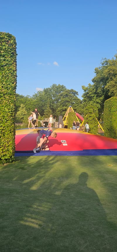 This image shows a vibrant outdoor playground scene in Groenlo, Netherlands, bathed in the warm glow of a sunny day. The focal point is a large, inflatable structure with sections in red, yellow, and blue, which appears to be a trampoline or bouncy castle. Several children are actively engaged on this inflatable surface, with some jumping and others interacting. In the foreground, a young boy in a blue and yellow jersey is leaning over, with another child sitting on the inflatable beneath him.
Behind the inflatable, a yellow, triangular climbing structure, possibly part of a larger play area, is visible, partially covered in greenery. The background is dominated by lush green trees and a clear blue sky. The ground surrounding the inflatable is covered in well-maintained green turf, with dappled sunlight and shadows creating a dynamic visual effect. Tall, manicured hedges frame the left and right sides of the scene, adding to the park-like atmosphere. The overall impression is one of joyful recreation and a bright, pleasant day spent outdoors.