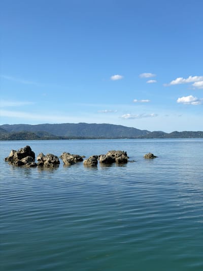 The image shows a tranquil seascape with a clear blue sky dotted with fluffy white clouds. The sea is calm and reflects the sky's blue, with gentle ripples disturbing the surface. A line of dark, rugged rocks emerges from the water, creating a natural barrier or perhaps a remnant of a past structure. The backdrop is a lush, green mountainous landscape that stretches along the horizon, hinting at a tropical or subtropical location. The sun appears to be high in the sky, suggesting it is daytime, likely midday or early afternoon, given the bright illumination and clear visibility. The overall scene conveys a sense of peace and natural beauty. The image is oriented vertically, with the sky on the left and the sea and land on the right.