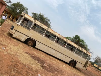 The scene, located in Ayangba, Nigeria, presents an outdoor environment under a light blue sky with scattered white clouds, viewed with an upward diagonal tilt. The ground consists of reddish-brown earth with patches of dry, light-colored vegetation and scattered white debris. A prominent, heavily weathered, light-colored (beige/off-white) bus is centrally positioned, appearing abandoned and partially sunken into the ground. The bus displays significant rust, peeling paint, and dirt accumulation on its exterior and roof. Most windows are missing or broken, exposing empty frames. Faint dark lettering, possibly "T.S.BOL," is visible on the side panel near the front. The bus's wheels are dark and appear to be either missing tires or severely deflated, submerged in the soil. One individual, wearing a white short-sleeved top and green trousers, stands on the far left of the frame near the front of the bus, facing left. In the background, a reddish-brown building with a dark roof is visible on the left, with additional smaller, lighter-colored buildings with dark roofs partially seen on the right. Trees with varying degrees of foliage are present in the mid-ground and background.