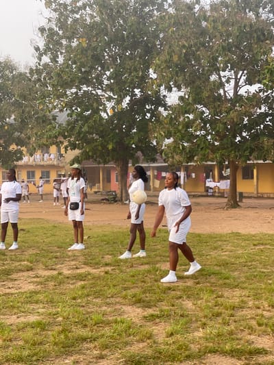 In Ise - Ekiti, Nigeria, a group of young women in white t-shirts and white shorts are engaged in an outdoor activity on a grassy field with dirt patches. The women are wearing white sneakers, and some have green and white striped socks. One woman in the foreground on the right is holding a light-colored ball. In the background, there are yellow and pink buildings with some laundry hanging outside. Several trees with green leaves are present on the field and in front of the buildings. The sky is overcast and appears hazy. A volleyball net is visible in the mid-ground.