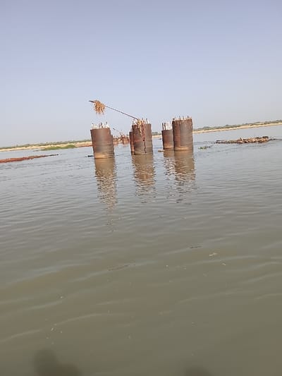The image depicts a wide, murky, greenish-brown body of water under a clear, pale blue sky. In the mid-ground, a series of at least eight large, rusty-brown cylindrical metal structures, likely caissons or coffer dams, are partially submerged, extending above the water surface. Reinforcing bars (rebar) protrude from the tops of several cylinders, indicating ongoing or abandoned concrete work. Accumulated organic debris, appearing as dried vegetation, is draped over the tops of some cylinders, and a cable or rope similarly laden with debris is suspended between two of these structures. Clusters of floating aquatic vegetation and dark, possibly wooden, debris are visible on the water surface. A distant, low-lying riverbank with sparse, dry-looking vegetation forms the horizon. No individuals are present. The scene is located in Ibi, Nigeria, consistent with a riverine environment.