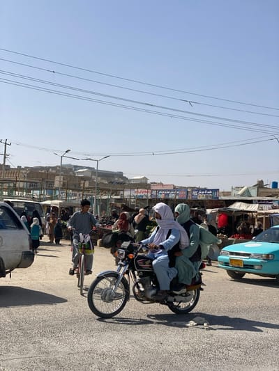 A motorcycle with several people crammed on is riding along a street. Other vehicles surround it and other people can be seen walking. Buildings, utility poles, and wires fill the background. The sky is a clear, light blue.