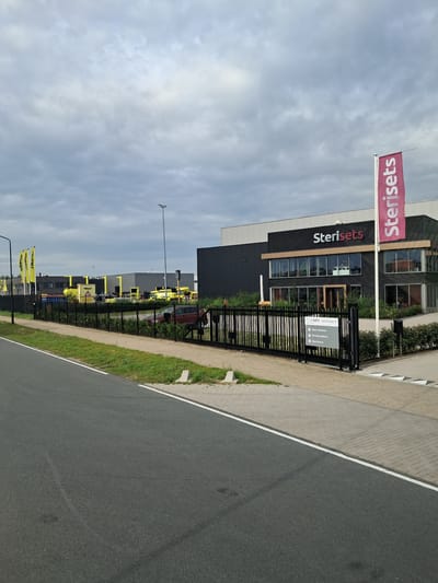 The image shows a modern industrial building with a prominent "Sterisets" logo and a vertical flag in front. The building is dark gray with a black facade and features large windows on the upper floor. In the foreground, a black metal fence encloses the property, with a sign near the gate indicating "Main entrance," "Parking visitors," and "Warehouse." To the left of the building, yellow flags with a logo are visible. Several yellow vehicles and industrial equipment are parked within the fenced area. The scene appears to be an office and warehouse facility for Sterisets, likely located in an industrial zone in Oss, Netherlands, given the presence of Dutch text on the sign. The sky is cloudy, suggesting overcast weather. The image is taken from a street level perspective, looking towards the entrance of the facility.