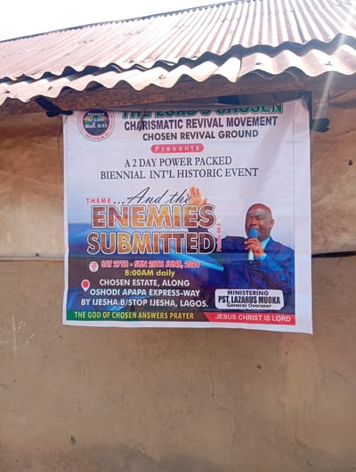 A wide shot shows a banner hanging on a rustic wall, presumably in Karsana 2, Nigeria. The banner announces a "2 DAY POWER PACKED BIENNIAL INT'L HISTORIC EVENT" by "THE LORD'S CHOSEN CHARISMATIC REVIVAL MOVEMENT," held at the "CHOSEN REVIVAL GROUND." The theme is "And the ENEMIES SUBMITTED," with the date "SAT 27TH - SUN 28TH JUNE, 2026" and time "8:00AM daily." The location is listed as "CHOSEN ESTATE, ALONG OSHODI APAPA EXPRESS-WAY BY IJESHA B/STOP IJESHA, LAGOS." A man in a suit, identified as "PST. LAZARUS MUOKA, General Overseer," is depicted holding a microphone. The banner also features biblical references, including "Heaven At Last Matt. 6:33" and "Psalms 66:3," and concludes with "JESUS CHRIST IS LORD." The background is a rough, textured wall and a corrugated metal roof.