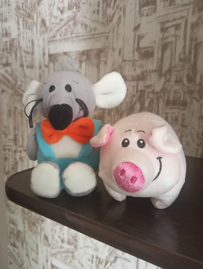 Two plush toys, a gray mouse wearing a blue shirt and orange bow tie and a pink pig, sit on a dark wooden shelf in front of a wall with a light brown and beige patterned wallpaper. The mouse is on the left and the pig is on the right. The scene is an indoor setting.