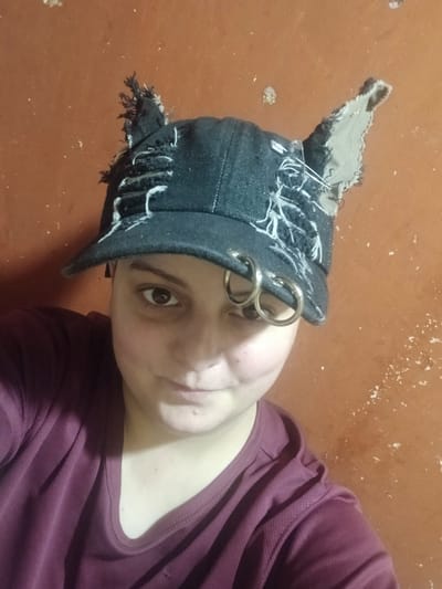 A person wearing a black denim baseball cap with cat-like ears made from ripped fabric and adorned with two large silver hoop earrings on the side of the cap is pictured. The person is looking towards the camera, with their right eye visible. They are wearing a dark maroon t-shirt. The background is a textured, orange-brown wall with some white specks. There are no identifiable landmarks or indications of a specific location within Chutove, Ukraine. The lighting is dim, and the image appears to be taken indoors.
