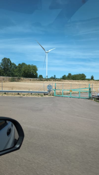 This is a vertically oriented photograph taken from a vehicle, likely during the daytime. The scene depicts a rural landscape with a clear blue sky featuring wispy clouds.
In the left portion of the image, a large white wind turbine stands tall against the sky, its blades extended. The ground around the turbine appears to be a field of dry, harvested crops, with scattered green trees visible further back.
To the right of the field, a bright green metal gate with a wire mesh fence separates the field from an asphalt road. A circular sign with a black border and a gray interior is mounted on a post near the gate. The road itself occupies the right side of the frame, showing a textured asphalt surface. A side mirror of the vehicle from which the photo is taken is visible in the lower right corner, reflecting part of the car's interior and the surrounding environment.
The presence of the wind turbine and the agricultural field suggests a rural setting, consistent with the provided location context of Sainte-Colombe, France. The clear sky and bright lighting indicate it is likely daytime, and the dry field suggests it might be late summer or early autumn. No people are visible in the image. There is some faint text on a yellow sign attached to the green gate, but it is not legible. The overall impression is one of open space and natural energy.