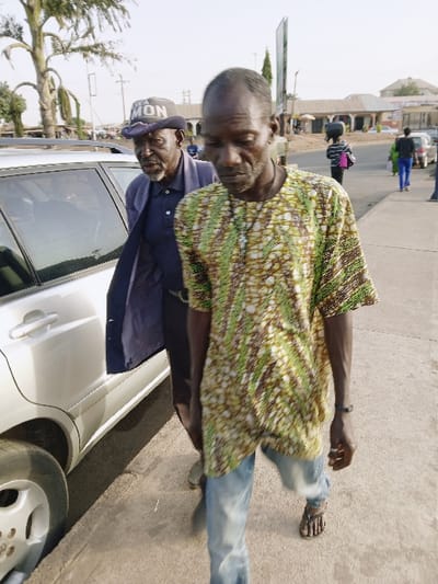 The image captures an outdoor street scene under bright daylight, understood to be in the vicinity of Kungulu School, Zambia. In the foreground, a thin, dark-skinned male, appearing middle-aged, walks towards the observer's left with his head lowered. He is dressed in a short-sleeved, intricately patterned shirt in shades of green, brown, and off-white, light blue jeans, and dark sandals. A light-colored cross pendant hangs from his neck. To his left and slightly behind him, an elderly dark-skinned male with a grey beard is positioned beside the open rear passenger door of a silver or light-colored SUV. He wears a dark blue long-sleeved garment over a darker shirt and a dark baseball cap with white lettering, partially visible as "MON". His face is turned towards the right, and his mouth is slightly agape.
The immediate ground consists of a dry, light-colored concrete or paved sidewalk. In the mid-ground and background, a roadway is visible, lined with various single-story and two-story buildings, some featuring reddish-brown roofs. Several individuals are present in the background: one pedestrian in a striped top carries an item on their head, walking away from the camera; others are seen further down the street. A large tree with sparse foliage is visible on the left, behind the parked SUV. A tall, dark vertical signpost stands in the mid-background on the right. Overhead utility lines are present against a clear, bright sky.
