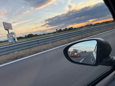 A side-view mirror of a vehicle reflects an orange hatchback car with a black hood driving on a highway. The car in the mirror has a license plate that reads "08 BB 2183". The highway is bordered by a metal guardrail and dry grass. In the distance, trees line the horizon under a sky with dramatic clouds illuminated by the setting sun. To the left of the highway, a billboard with the word "ELIT" is visible. The image was taken on the highway near Kostievo, Bulgaria.