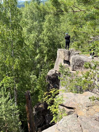 A person wearing a black t-shirt and black pants stands on a rocky cliff overlooking a dense forest. The person's back is to the camera, and they appear to be looking out at the landscape. The cliff is made of large, grey rocks, and there are green trees and vegetation surrounding it. The sky is visible in the background, and it appears to be a sunny day. This scene is located in Orlenok, Russia, specifically at the "Eagle Rock" tourist spot.