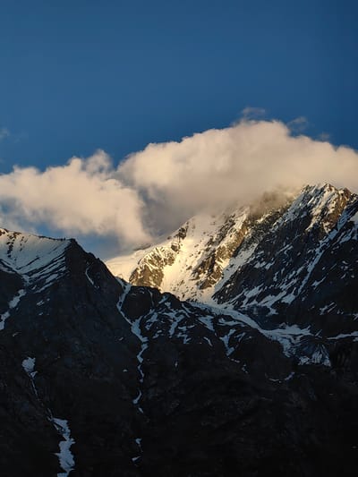 This media file depicts a majestic mountain landscape at sunrise in Gemur, India. The scene is dominated by rugged, snow-capped peaks bathed in the warm, golden light of the rising sun. A large, fluffy cloud formation hovers above the mountains, with some parts catching the sunlight and appearing luminous. The sky is a deep, clear blue, contrasting with the snowy white and dark gray of the mountains.
The foreground is cast in shadow, highlighting the dramatic contours and textures of the mountainside. Patches of snow cling to the steep slopes, interspersed with exposed rock faces. The lighting suggests the early morning, with the sun just beginning to illuminate the higher elevations. The weather appears clear with some cloud cover at higher altitudes.
While there are no distinct objects or people visible, the grandeur of the natural setting evokes a sense of awe and tranquility. The image captures the raw beauty of the Himalayas, with the play of light and shadow creating a dramatic and captivating scene. The location context of Gemur, India, places this dramatic sunrise within a region known for its stunning mountain vistas.