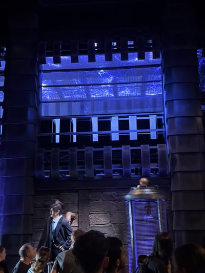 The image captures a scene with a theatrical or exhibit-like atmosphere, predominantly lit with deep blue and purple hues. The setting appears to be indoors, possibly a themed attraction or performance space, with a dark, industrial-inspired backdrop. Towering, dark, segmented structures frame the scene on either side. In the background, a large illuminated panel displays a bright blue, abstract pattern resembling electrical discharges or veins.
In the foreground, a group of people are visible, their faces mostly obscured as they look towards a central figure. The central figure is a person dressed in a dark suit with a white shirt and tie, appearing to be on a stage or platform. To the right of the central figure, a tall, cylindrical structure with a caged exterior and a golden spherical ornament on top emits a blue glow. The overall ambiance is dramatic and visually striking, with a focus on artificial lighting and a sense of mystery or spectacle. The image was taken in Rust, Germany.