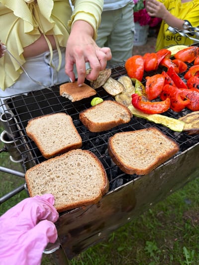 A close-up, high-angle shot captures a grill laden with food, including slices of bread, grilled red bell peppers, zucchini, and eggplant. The scene is set outdoors, with green grass visible in the foreground and background. In the upper left, a person wearing a light yellow, ruffled top and white pants is seen holding a fork and reaching towards the grill. Their hands are positioned over some of the grilled food. To the right, another person, partially visible, wears a bright yellow t-shirt with "SHG" printed on it. In the lower left, a pink gloved hand is visible, touching the edge of the grill. The overall impression is of a casual outdoor gathering and food preparation, likely taking place in a park or backyard. The specific location of Bronnitsy, Russia is not visually indicated in the image.