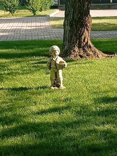 A statue of a young boy holding a bucket stands in a grassy area in the city of Pokrov, Ukraine. The statue is made of stone and appears to be weathered. The boy is wearing a smock and shorts, and his hair is curly. He is standing on a paved walkway in the background, and there are trees and bushes around. The sun is shining, and the grass is green and well-maintained. The overall scene is peaceful and serene, suggesting a park or garden setting.