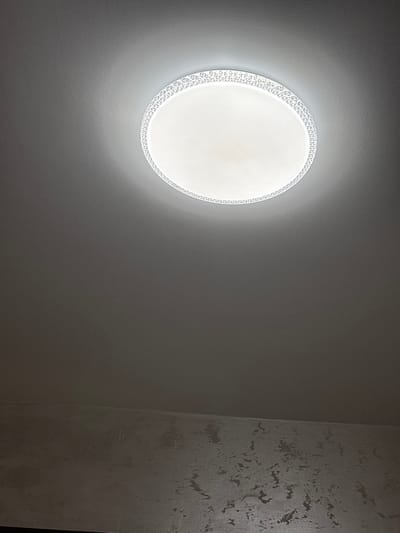 The image depicts an interior architectural space located in the city of Bukovlak, Bulgaria. A circular, surface-mounted LED light fixture is affixed to a flat, light grey ceiling, positioned in the upper-center of the frame. The fixture emits white light, illuminating the surrounding ceiling and an adjacent wall. The light fixture features a central translucent diffuser and a perimeter ring with a repeating geometric pattern. The wall, visible in the lower portion of the image, exhibits an irregular, mottled grey texture, distinct from the smoother ceiling surface. No human subjects, objects other than the fixture, or discernible actions are present.