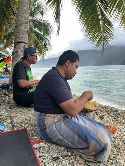 Two men are sitting on a pebbly beach next to a palm tree, facing the ocean. The man on the left is wearing a black t-shirt, a blue baseball cap, and glasses. He is looking out at the water. The man on the right is wearing a dark blue polo shirt and a patterned sarong. He is holding a clear plastic bowl filled with food, appearing to be noodles, and is in the process of eating. The ocean is a turquoise color with waves breaking on the shore. In the background, there are misty mountains. The environment appears to be tropical. The location is the city of Ujongsedheun, Indonesia.